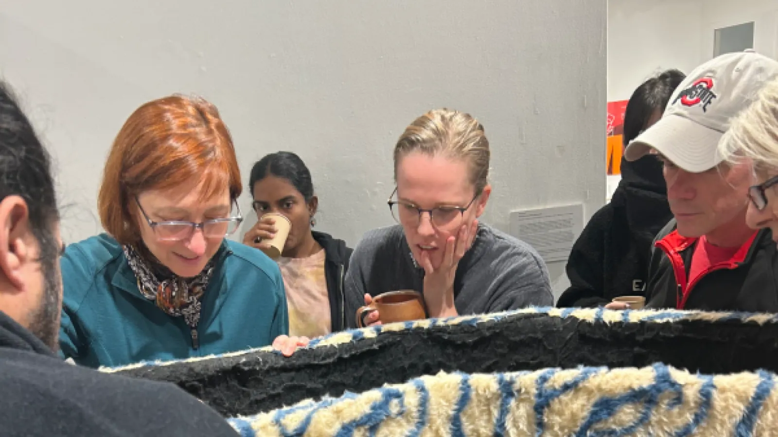 Close up of 7 Adults looking down into an open fuzzy container. The container is white with blue zebra stripes from the outside. Three figures hold cups or tea mugs. 
