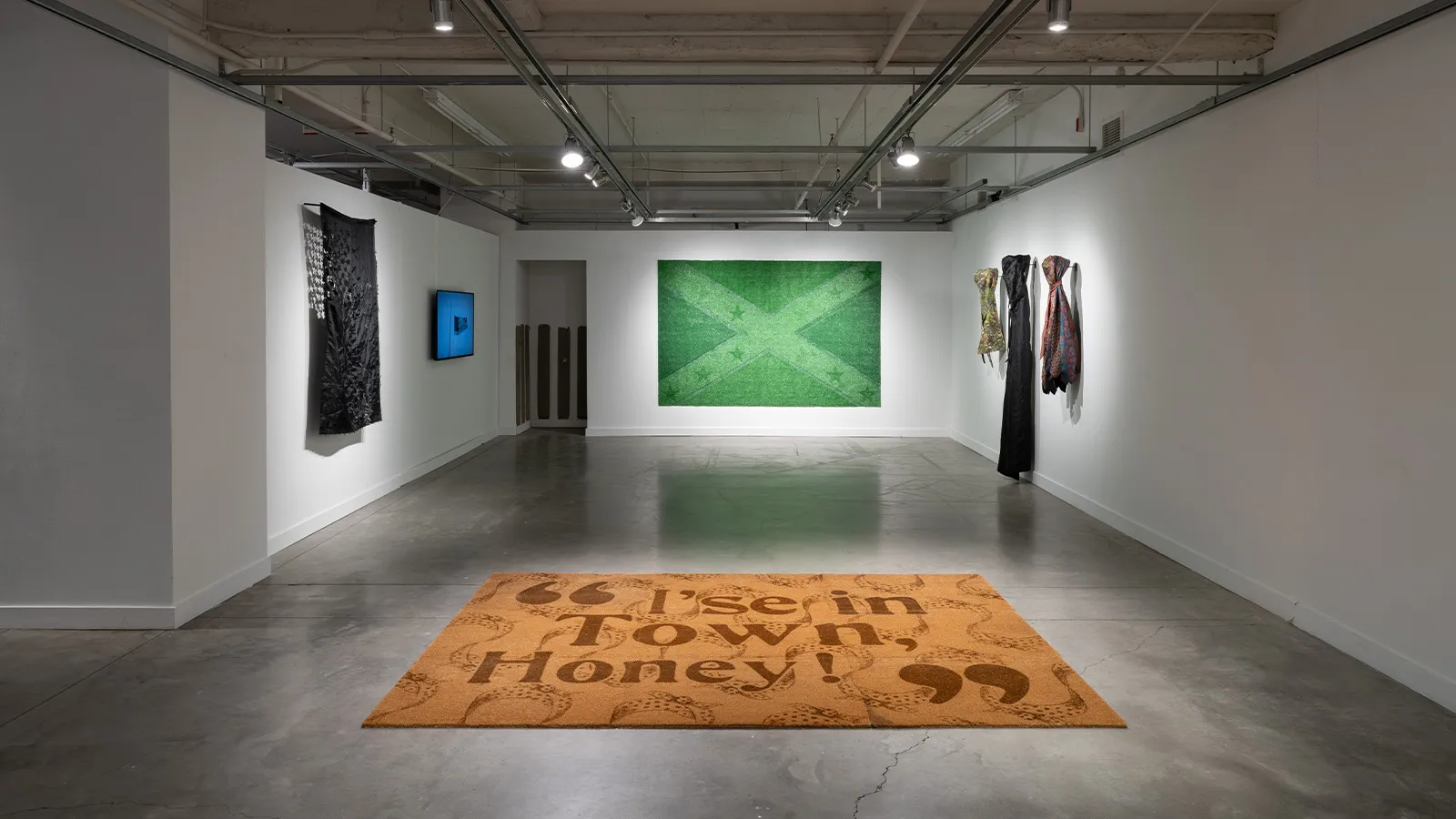 Installation view of artwork on floor and walls with flags, video, and welcome mat that reads, “I’se in Town Honey!”.  