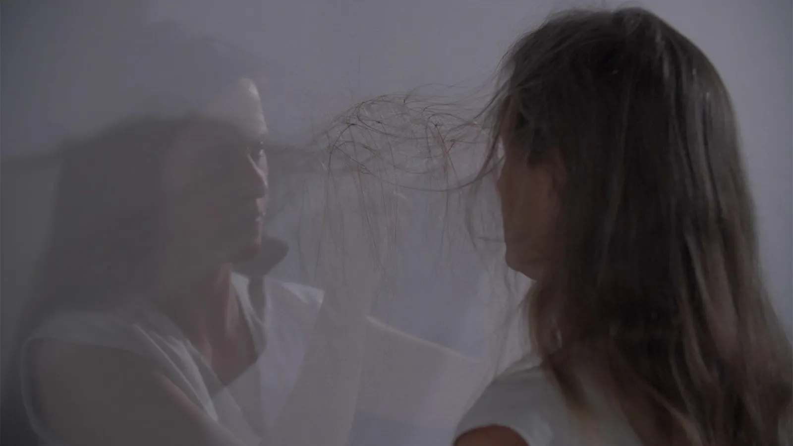 Still from a video performance of two women on opposite sides of a screen who are tying their hair together. 