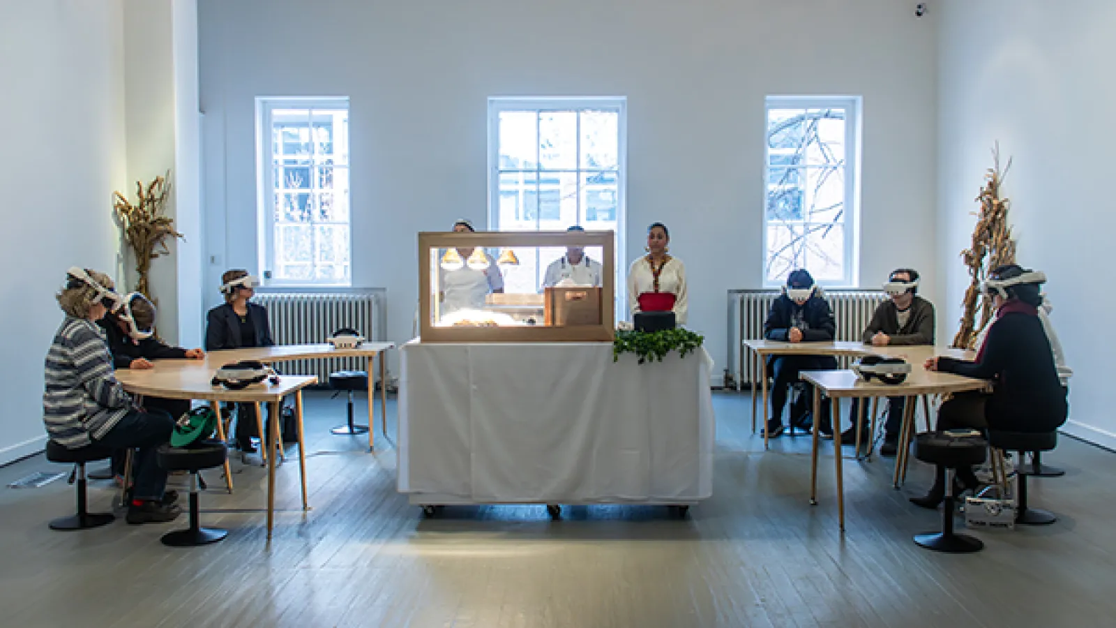 Visitors wearing VR headsets sit in a circular dining arrangement in a well lit gallery room.