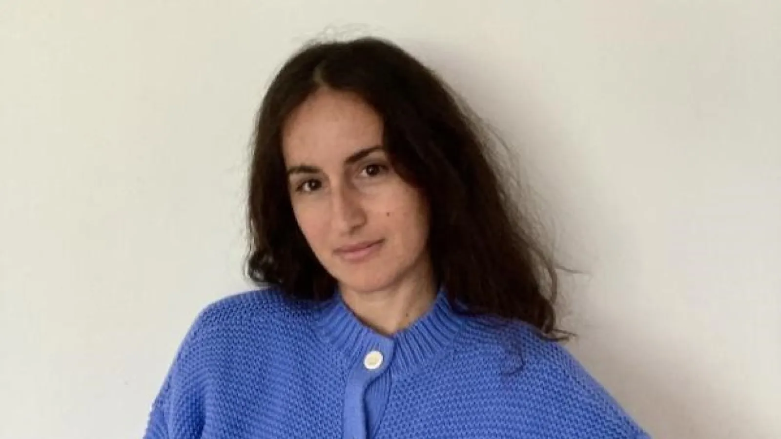 A medium close-up portrait of artist Carmen Winant, looking toward the camera against a plain, light-colored background. She has long, dark brown hair parted down the middle and is wearing a textured, cornflower blue knit cardigan with visible buttons at the collar.