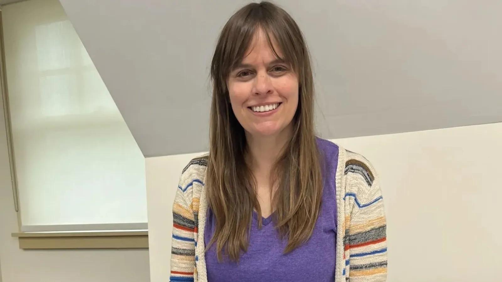 A close-up portrait of artist and professor Kate Shannon smiling warmly. She has long, straight brown hair with full bangs and is wearing a purple top underneath a cream-colored, open-front cardigan with vibrant multi-colored stripes on the cuffs and trim. She is standing indoors in front of a white wall.