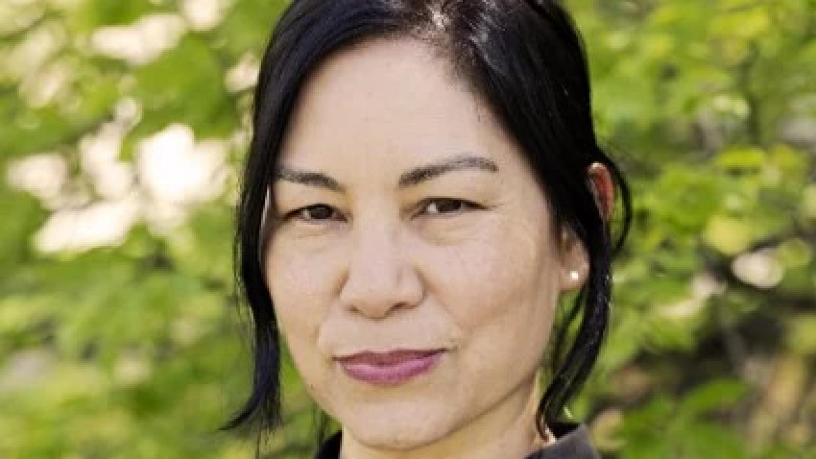 Portrait of Gina Osterloh, a person with dark hair wearing a black collared shirt, photographed outdoors against a softly blurred green leafy background