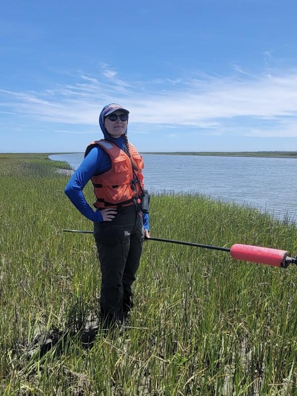 a person with an orange life vest, baseball cap and sunglasses standing in marsh grass