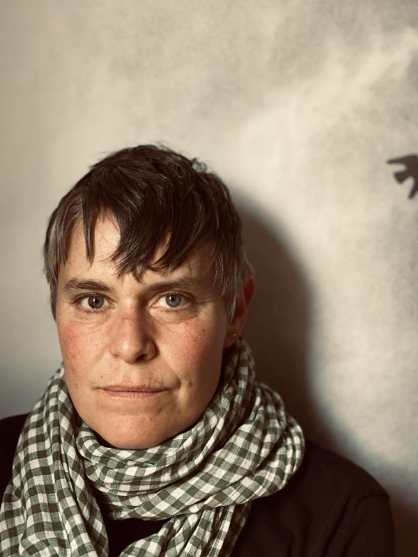 a person staring into the camera with a black and white scarf around their throat.  A single black bird is in silhouette behind them on the right. 