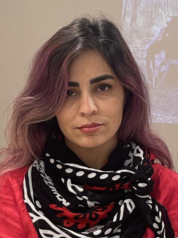 A person with long dark hair, dark eyes wearing a red shirt and black, white and red scarf